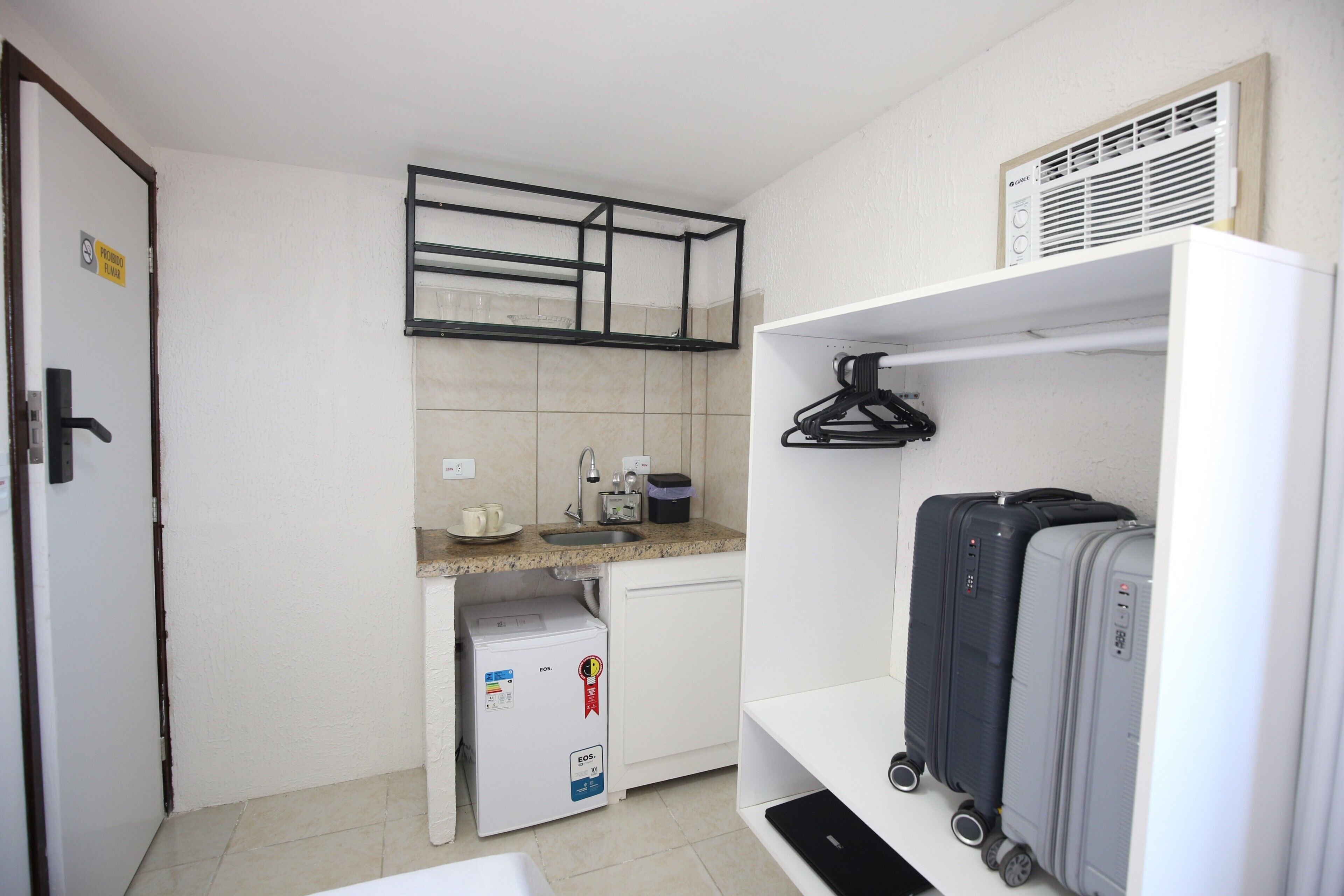 Una kitchenette compacta con refrigerador, pileta y una pequeña mesada. Hay dos maletas en un estante y una puerta con un cartel.