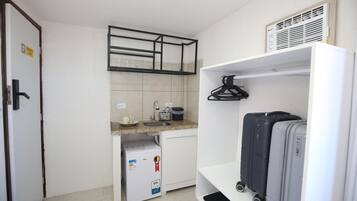 Una kitchenette compacta con refrigerador, pileta y una pequeña mesada. Hay dos maletas en un estante y una puerta con un cartel.