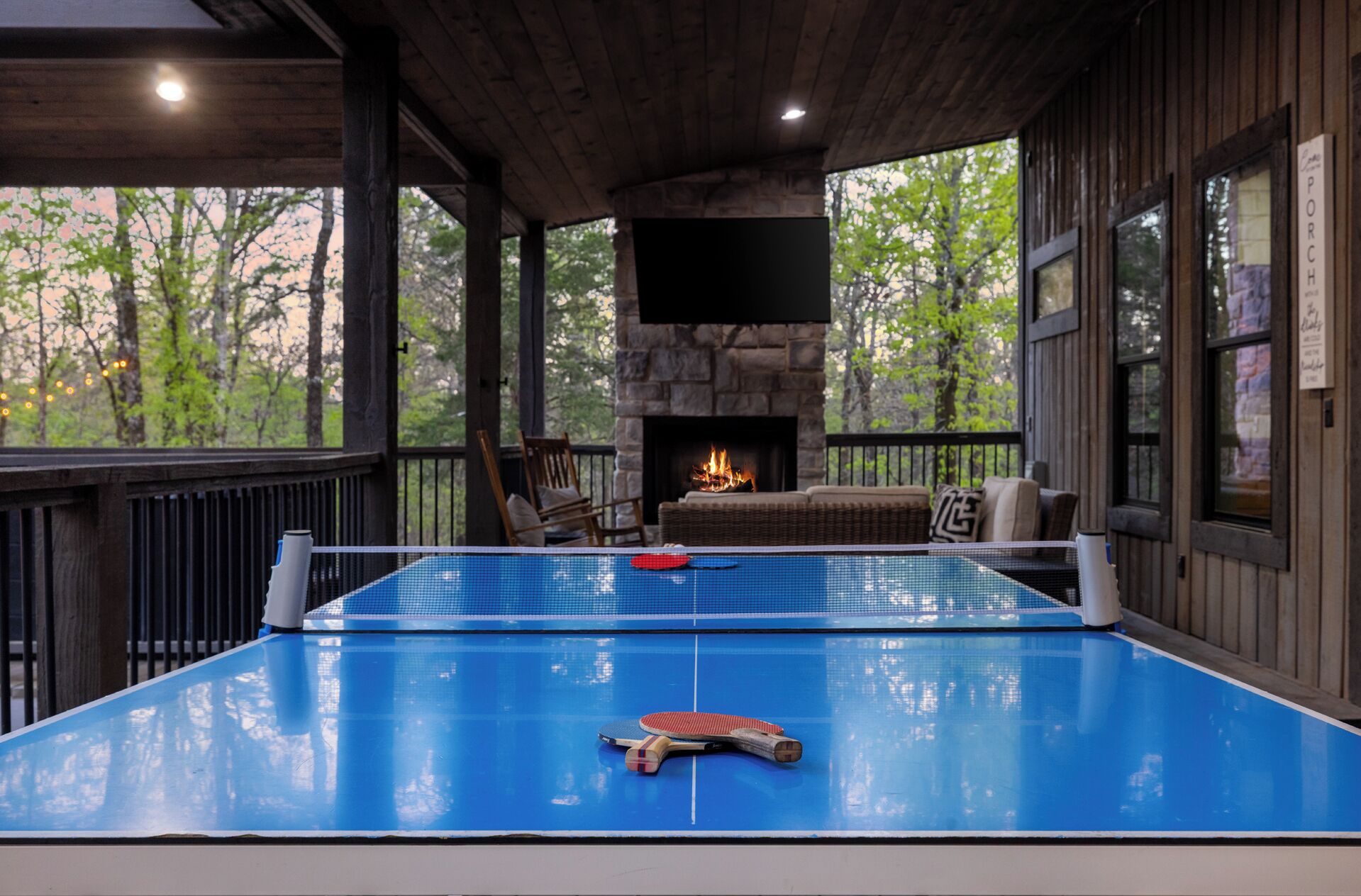 A ping pong table with paddles, a fireplace, and a wicker sofa on a porch with a view of trees.