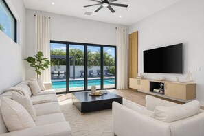 Ein modernes Wohnzimmer mit Blick auf einen Pool, weißen Möbeln und einem Flachbildfernseher.