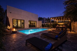 Ein moderner Poolbereich im Freien mit Liegebereich, einer Pergola mit Lichterketten und einem Feuerbereich.