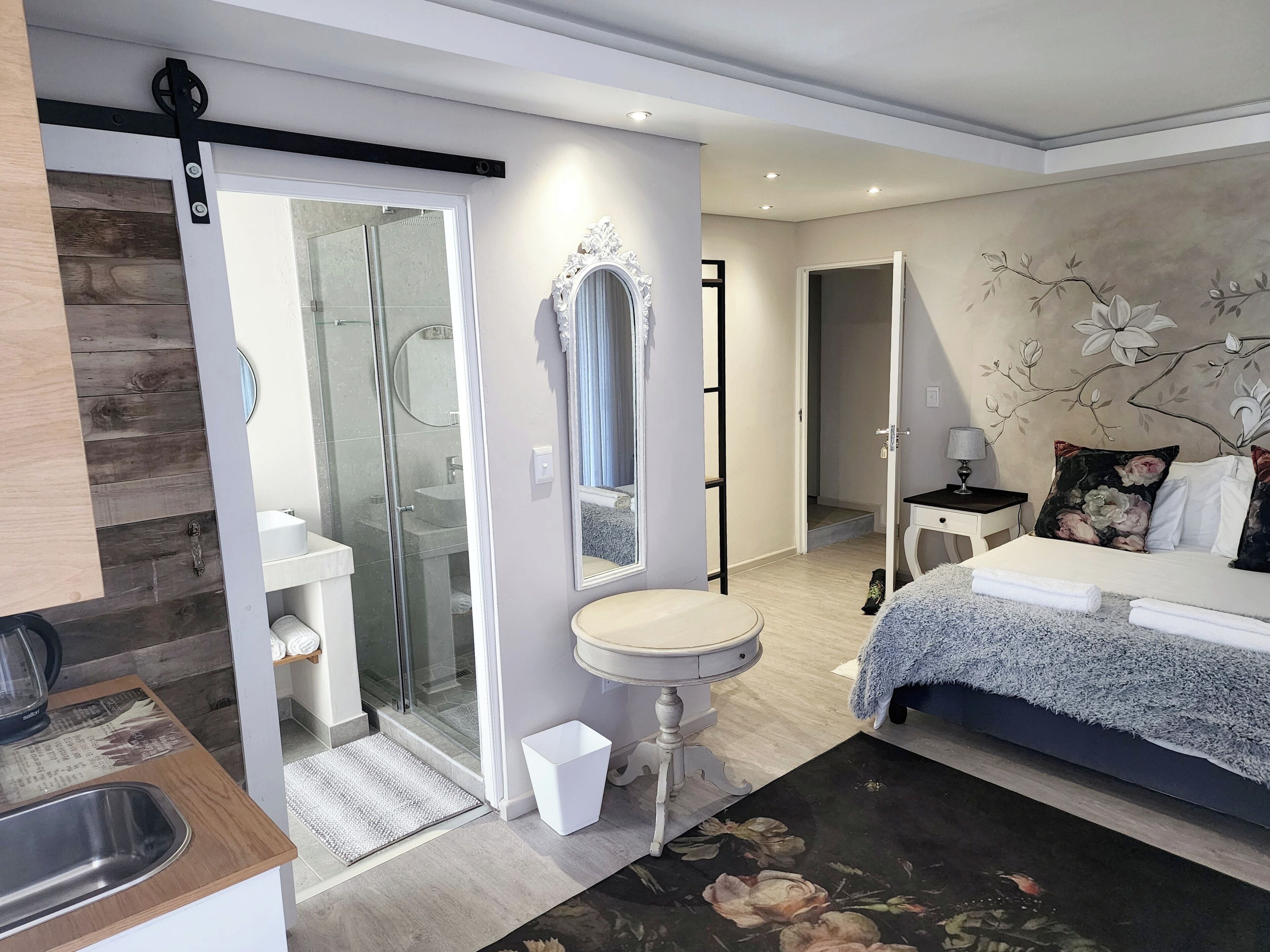 Une chambre moderne avec une douche, un miroir rond, une table de chevet avec une lampe, un lit agrémenté d’un coussin à motifs floraux et une vue sur le couloir.