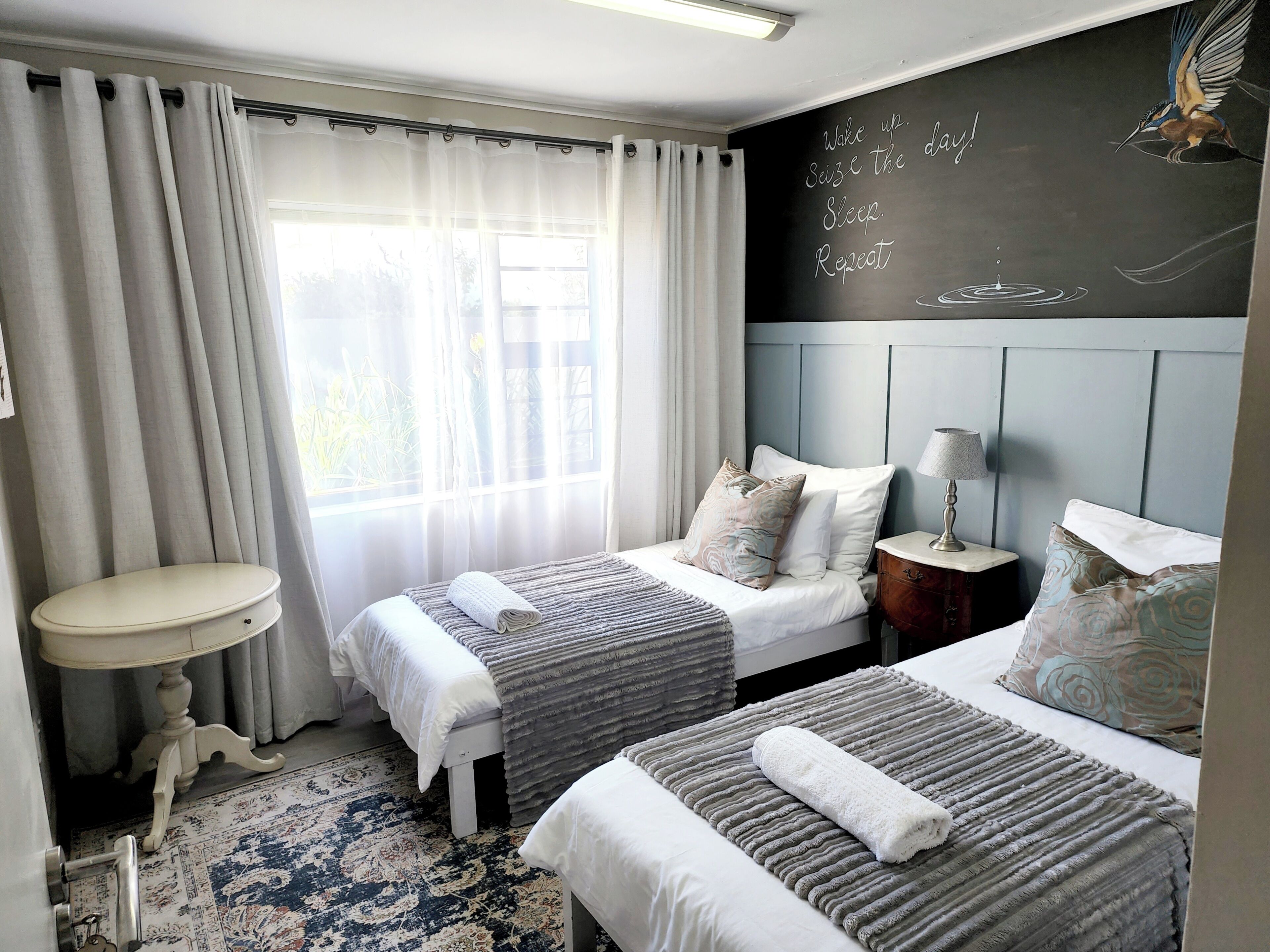 A hotel room with two beds, a small table, a lamp, and a chalkboard with motivational writing.