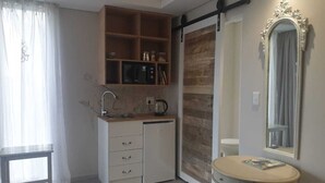 Una cocina compacta con un lavamanos, un refrigerador pequeño, un microondas y una cafetera. Hay un estante de madera sobre el lavamanos y un espejo también sobre el lavamanos.