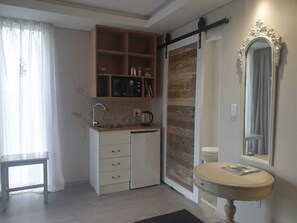 A compact kitchen with a sink, a small refrigerator, a microwave, and a coffee maker. There is a wooden shelf above the sink and a mirror above the sink.