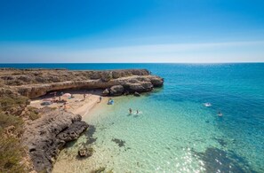 Ein Küstenstrand mit klarem Wasser, Felsformationen und Menschen, die die Sonne genießen.