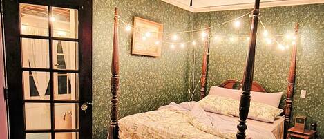 Un dormitorio con cama con dosel, ventilador de techo, guirnaldas de luces, una puerta con paneles de vidrio y una alfombra estampada.