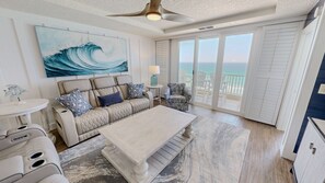 A spacious living area with a large window overlooking the ocean, a ceiling fan, and a painting of a wave. - The Inn At Crystal Beach Ph 702 4 Bedroom Condo (Destin)