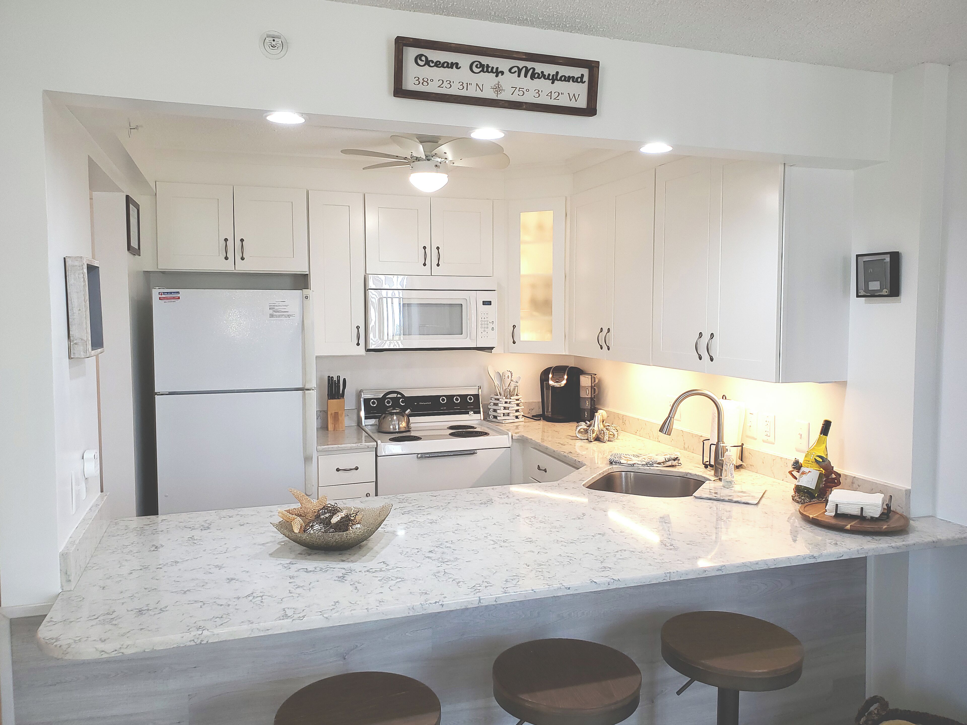 A kitchen with white cabinets, a refrigerator, a microwave, a stove, and a sink. There is a ceiling fan, a sign indicating the location, and a bowl on the counter.