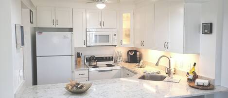 A kitchen with white cabinets, a refrigerator, a microwave, a stove, and a sink. There is a ceiling fan, a sign indicating the location, and a bowl on the counter.