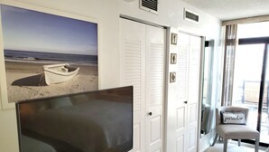 Une pièce avec un téléviseur à écran plat posé sur un meuble en bois, des portes blanches, un tableau représentant une plage et un fauteuil avec un coussin.