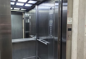 A metallic elevator interior with a control panel, emergency exit sign, and a door handle. - Palermo: Stylish Deco in a Functional Studio with Balcony (Buenos Aires)