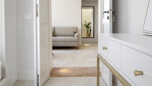 Ein Flur mit einer weißen Tür, einem beigen Sofa, einem Spiegel und einem weißen Schminktisch mit goldenen Griffen.