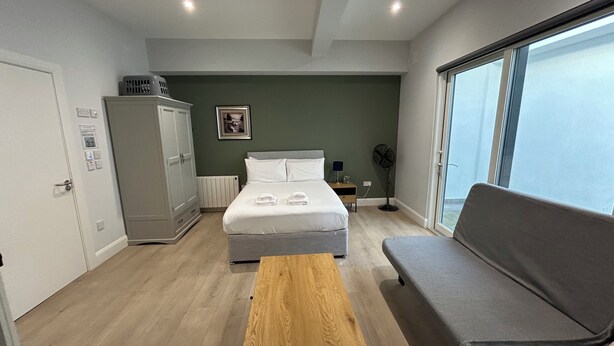 Une chambre à coucher avec un lit, une table en bois, un canapé gris, une armoire, un radiateur, un tableau accroché au mur et une fenêtre.