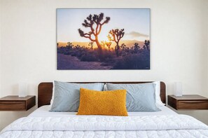 Ein Schlafzimmer mit einem Bett, zwei Nachttischen, einem an der Wand hängenden Bild einer Wüstenlandschaft und einem gelben Kopfkissen.