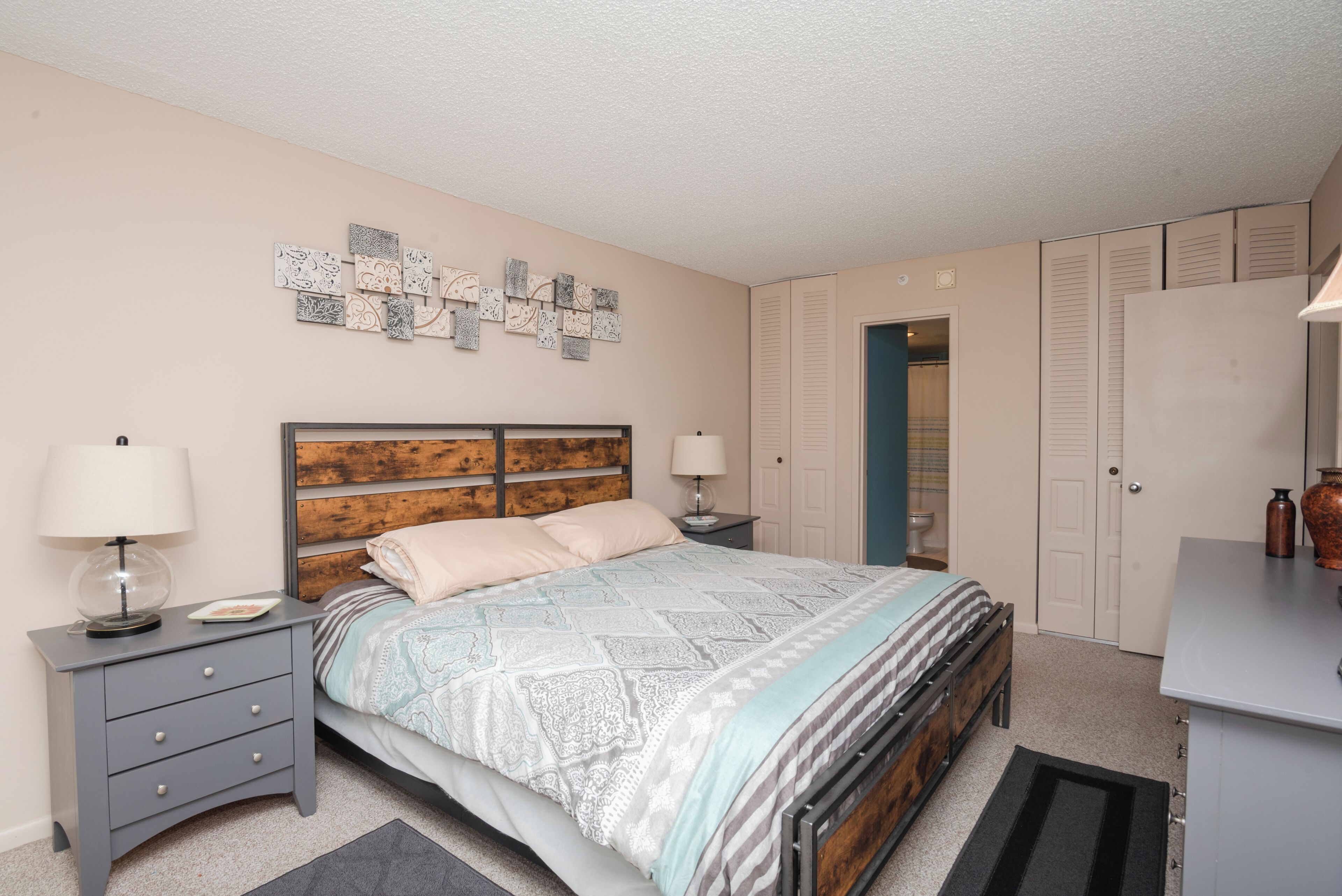 A bedroom with a wooden headboard, a bedside table with a lamp, a bed with patterned bedding, a closet with louvered doors, and a wall-mounted artwork.