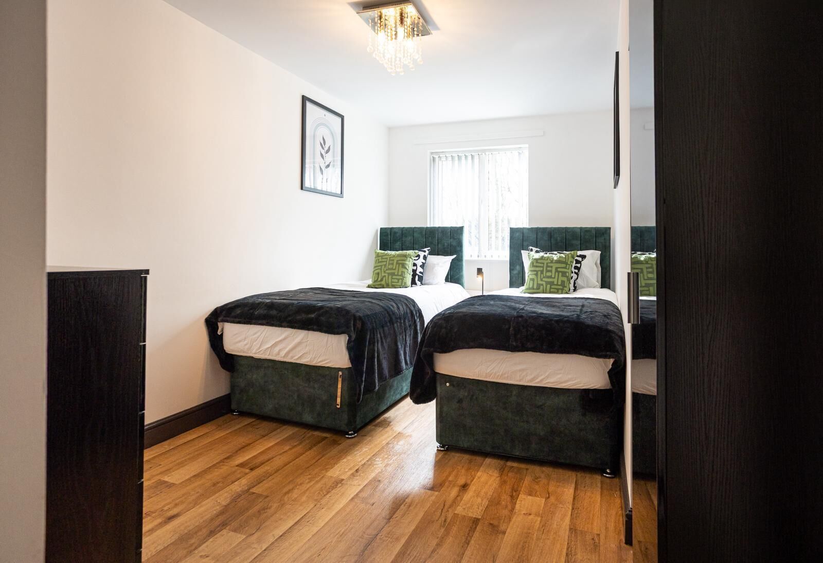 A room with two beds, a wooden floor, a framed picture on the wall, and a window with curtains.