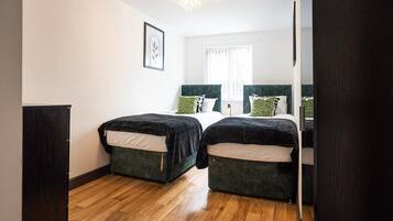 A room with two beds, a wooden floor, a framed picture on the wall, and a window with curtains.