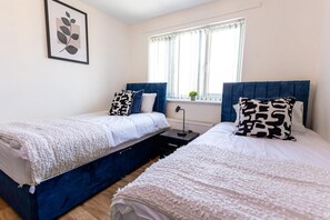 Une chambre à deux lits avec deux lits simples, une table de chevet avec une lampe, une fenêtre avec des rideaux et un tableau encadré au mur.