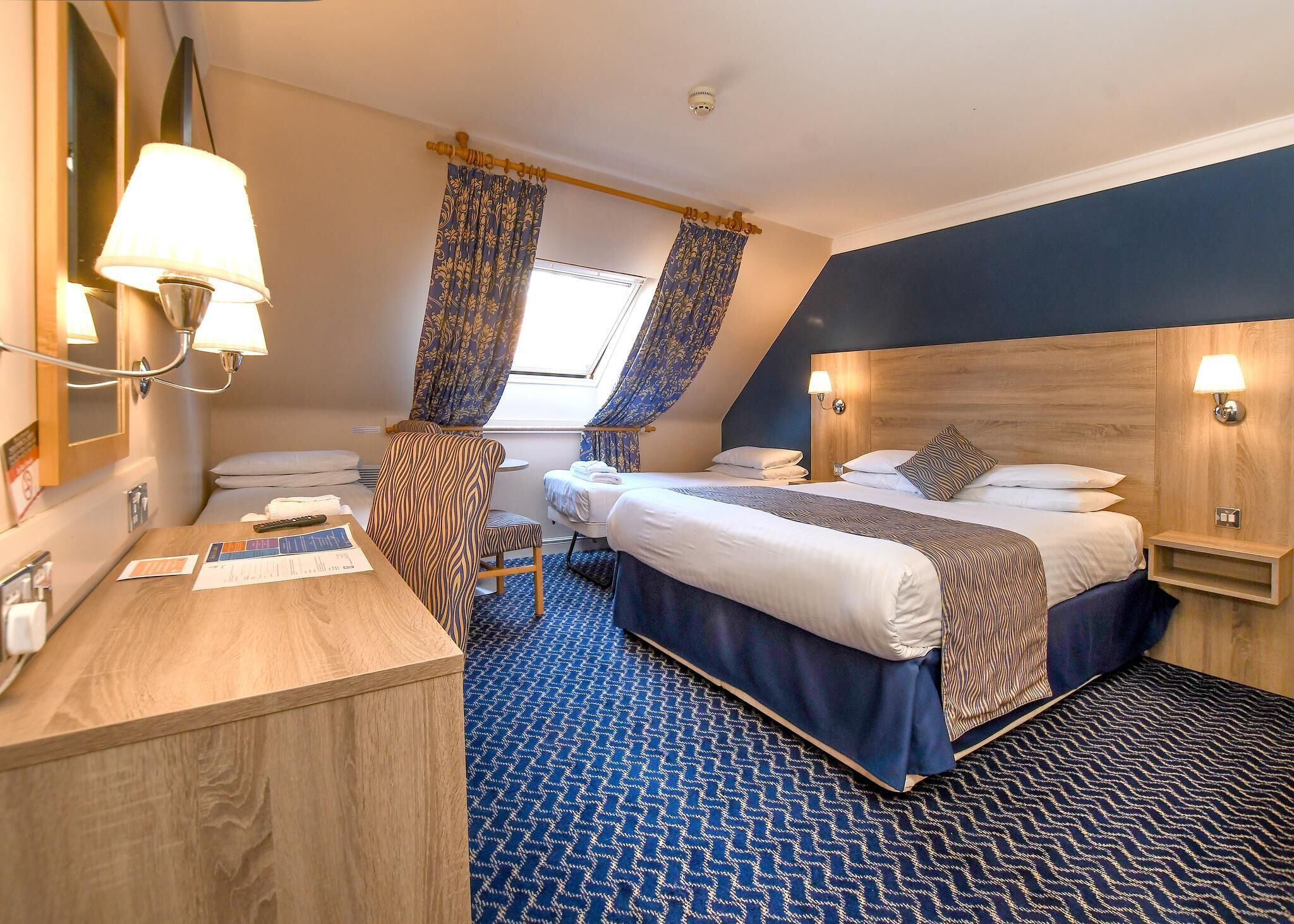 Ein Hotelzimmer mit zwei Betten, einem Schreibtisch, einem Stuhl, einem Fenster mit Vorhängen und einer Deckenleuchte.