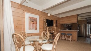 Ein Zimmer mit Holzwänden, einem Esstisch mit Glasplatte, umgeben von Korbstühlen, einer Küchenzeile mit Edelstahlgeräten und einem an der Wand befestigten Flachbildfernseher.