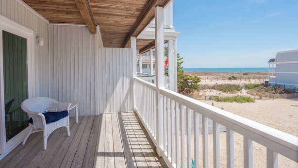 Eine Holzterrasse mit einem weißen Stuhl, Blick auf den Strand und einem Gebäude im Hintergrund.