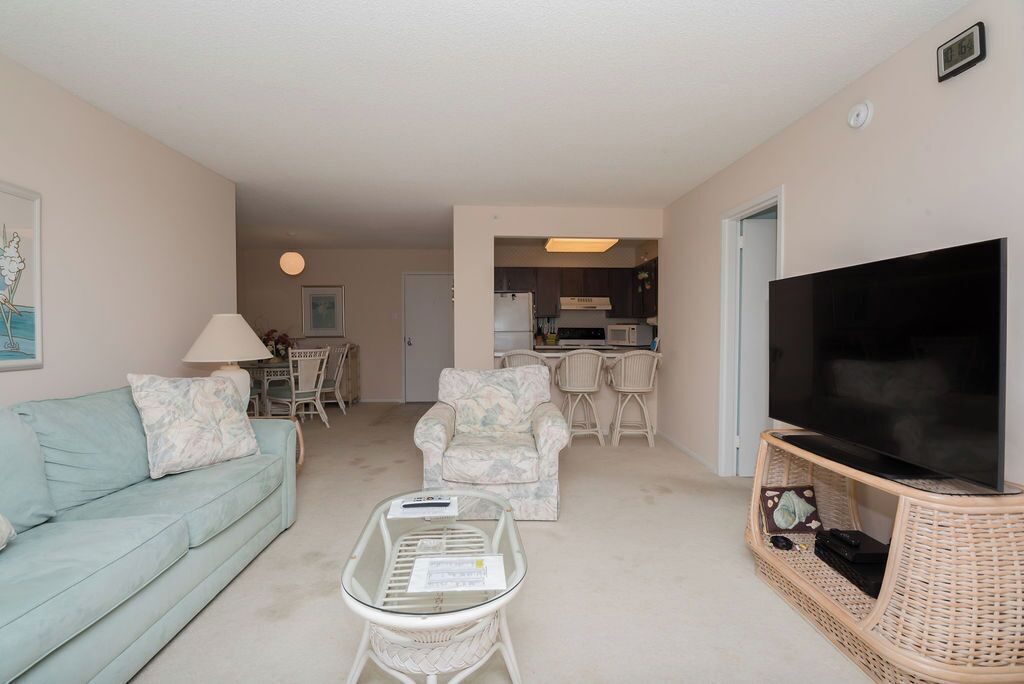 A living room with a sofa, armchair, coffee table, TV, and kitchen area in the background.