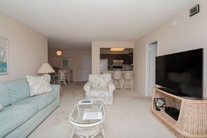 Ein Wohnzimmer mit Sofa, Sessel, Couchtisch, Fernseher und einer Küche im Hintergrund.