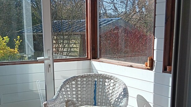 A covered porch with wicker chairs, a red cushion, and a view of trees and a building. - La Parenthèse enchantée (Le Vast)