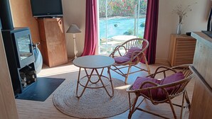 A room with a TV, a fireplace, wicker chairs, a table, and a view of a pool. - La Parenthèse enchantée (Le Vast)