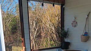 A covered patio with a table and chairs, a shelf with a basket, and a view of trees. - La Parenthèse enchantée (Le Vast)