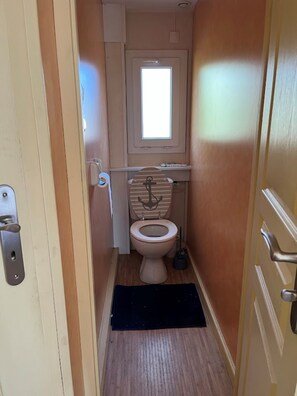 Een smalle badkamer met toilet, een klein raam en een deur met een hendel.