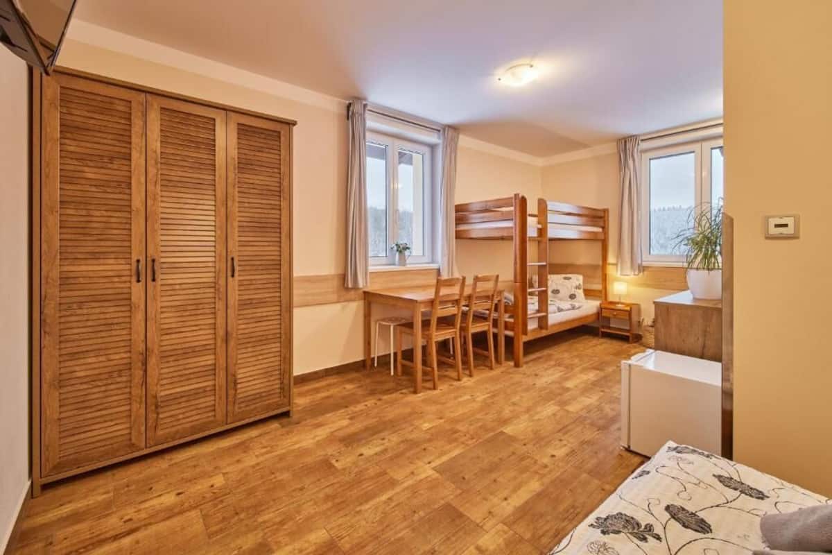 A compact room with a bunk bed, wooden wardrobe, a desk with chairs, a small table, and a window.