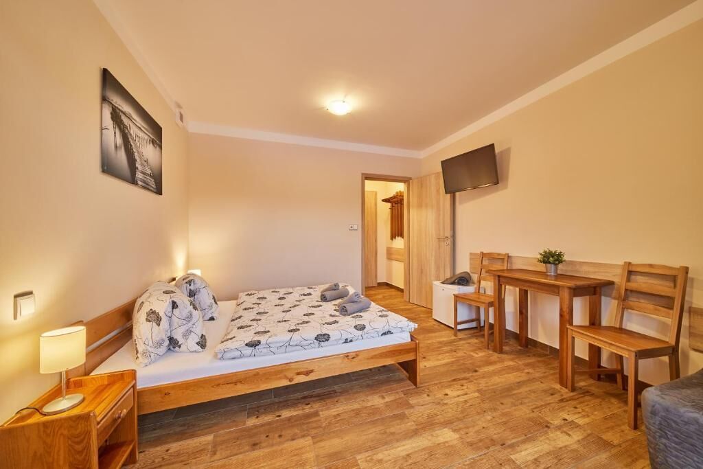 A bedroom with a bed, bedside table, lamp, TV, wooden desk, chairs, and a small plant.