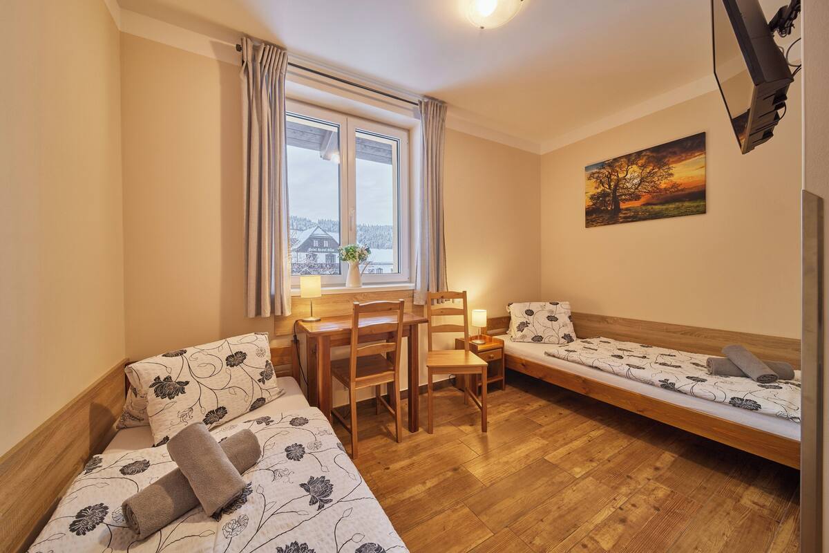 A room with two beds, a desk, chairs, a window with curtains, a painting, and a television.