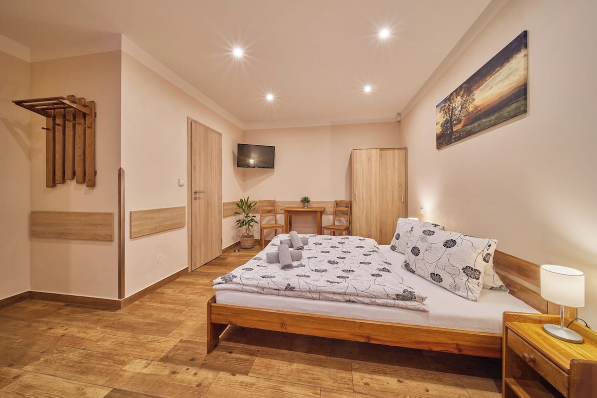 A bedroom with a wooden bed, a TV mounted on the wall, a desk with a chair, and a wooden wardrobe.