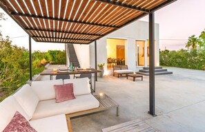 Um pátio externo com pérgola de madeira, um sofá, uma mesa de jantar posta para dois e uma escadaria que leva a um espaço interior.