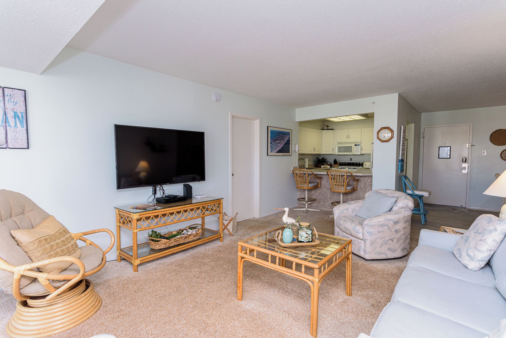 A living room with a television, wicker furniture, a white sofa, and a kitchen in the background.