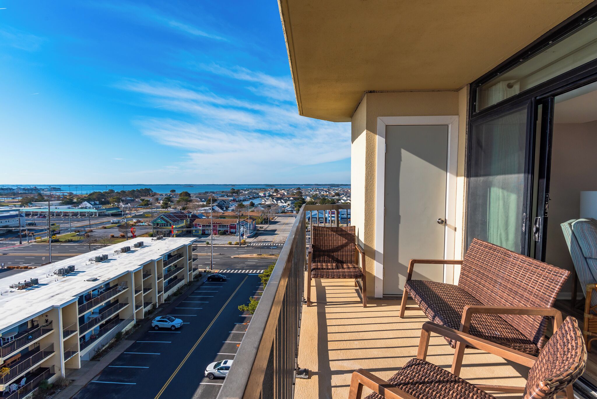 A balcony with wicker furniture, a view of a parking garage, and a distant cityscape.