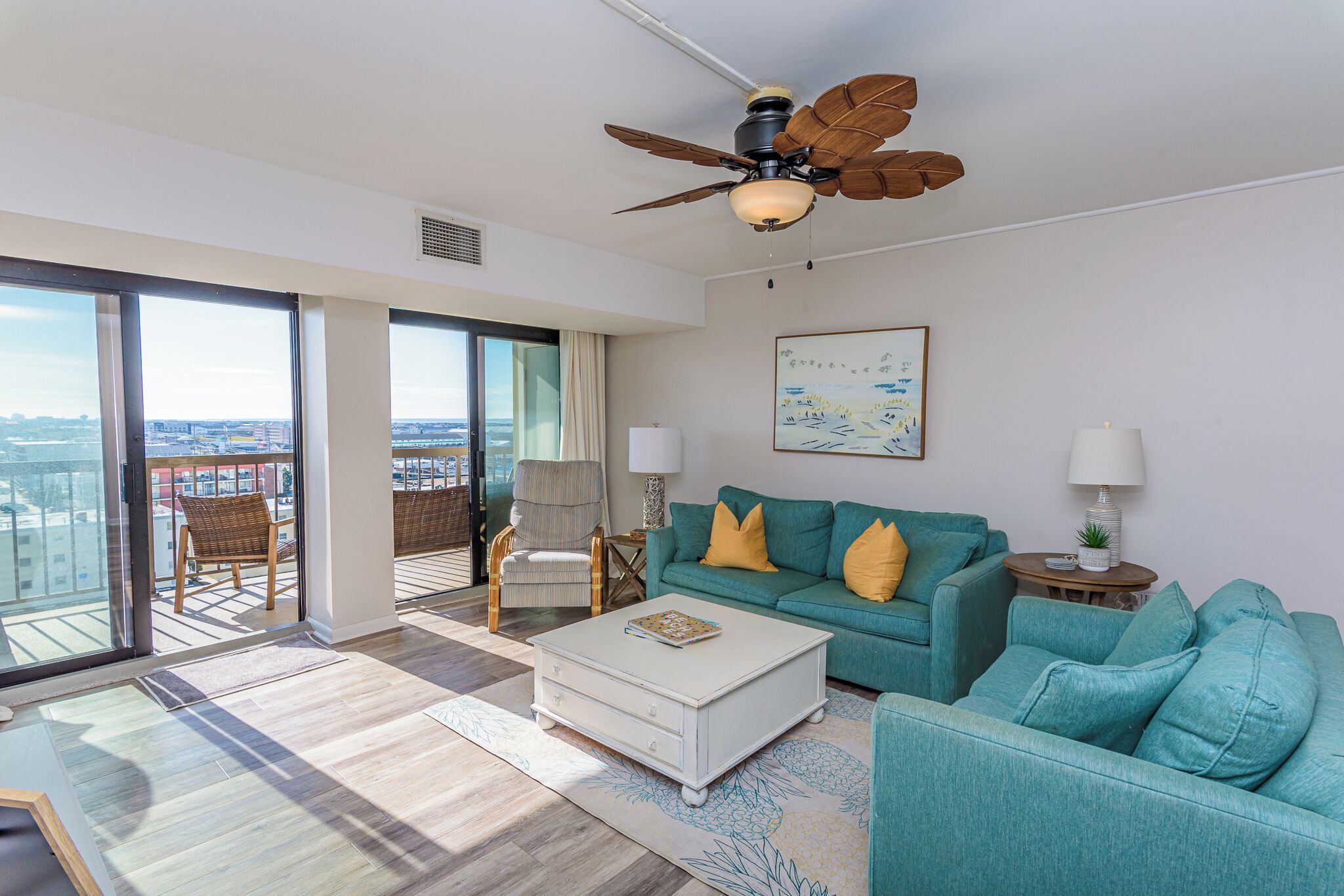 A spacious living room with a large window, a ceiling fan, a teal sofa, and a white coffee table.