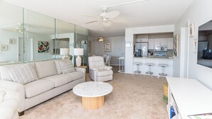 Een moderne woonkamer met een bank, fauteuil, en salontafel, een keuken met een magnetron en barstoelen, en een plafondventilator.