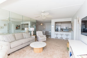 A modern living room with a sofa, armchair, and coffee table, a kitchen area with a microwave and bar stools, and a ceiling fan. - Oceanfront Building With Great Ocean and Bay Views (Ocean City)