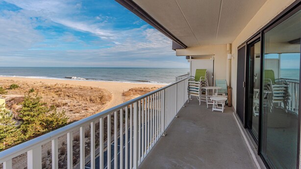 A balcony with a view of the beach, outdoor seating, and a glass door.