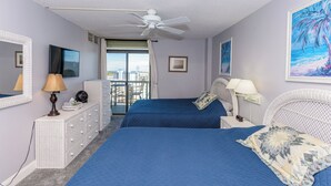 Un dormitorio con dos camas, ventilador de techo, televisor, cómoda y balcón con vistas.