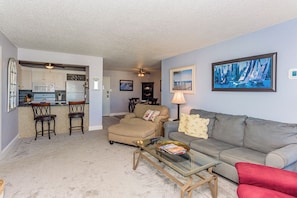 A living room with a sofa, a glass coffee table, a red chair, a kitchen area with a refrigerator, and a ceiling fan. - Oceanfront Building With Great Ocean and Bay Views (Ocean City)