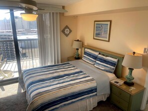Una camera d'albergo con un letto, un comodino, una lampada, un quadro, una finestra con tende e vista sugli edifici.