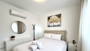 A neatly made bed with rolled towels, a round mirror, a wall-mounted air conditioner, a floor lamp, and a painting of a tree. - 127 NIC - Stay Sun Line Apartment (Nicosia)