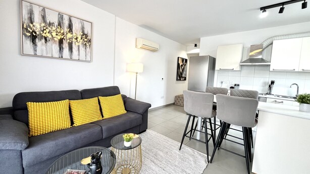A modern living room with a grey sofa, yellow cushions, a glass coffee table, and a kitchen area with white cabinets and a stainless steel range hood. - 127 NIC - Stay Sun Line Apartment (Nicosia)
