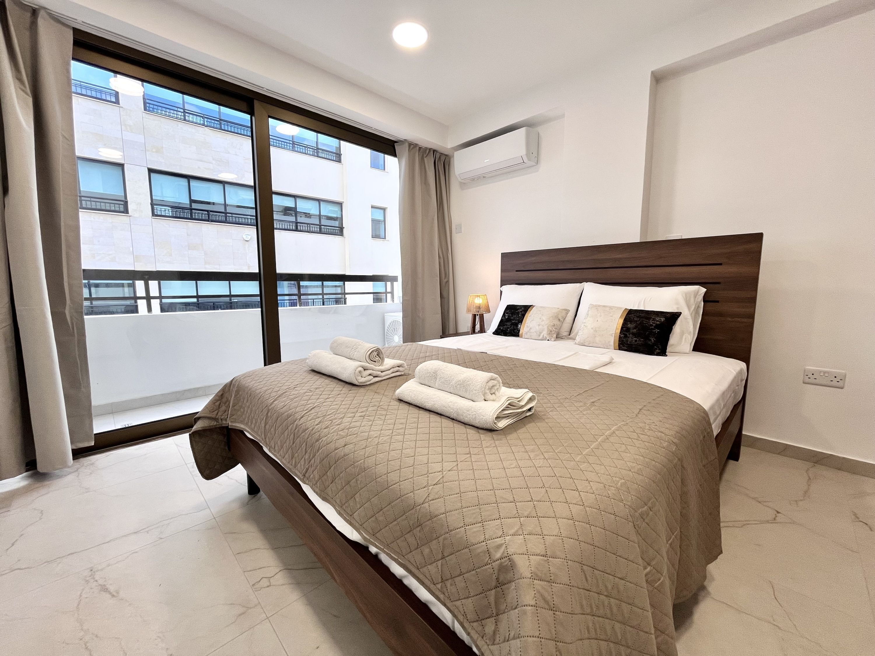 Quarto de hotel com uma cama grande, uma janela com vista para edifícios, um ar condicionado e uma mesa de cabeceira com abajur.