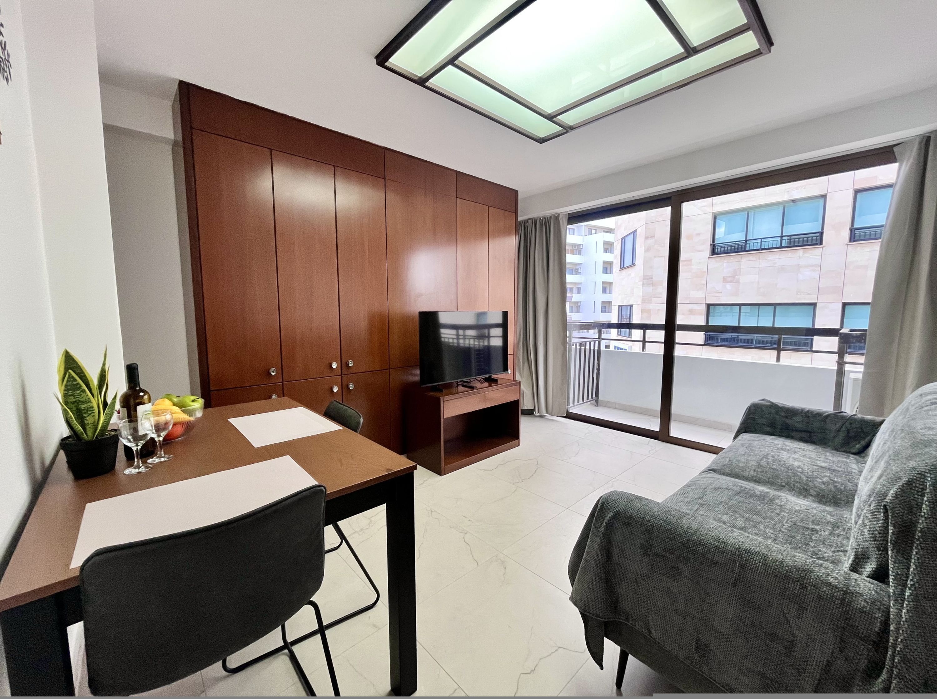 Uma sala de estar moderna com um armário de madeira, uma televisão de tela plana, uma mesa de jantar com cadeiras, um sofá e uma varanda com vista para a cidade.